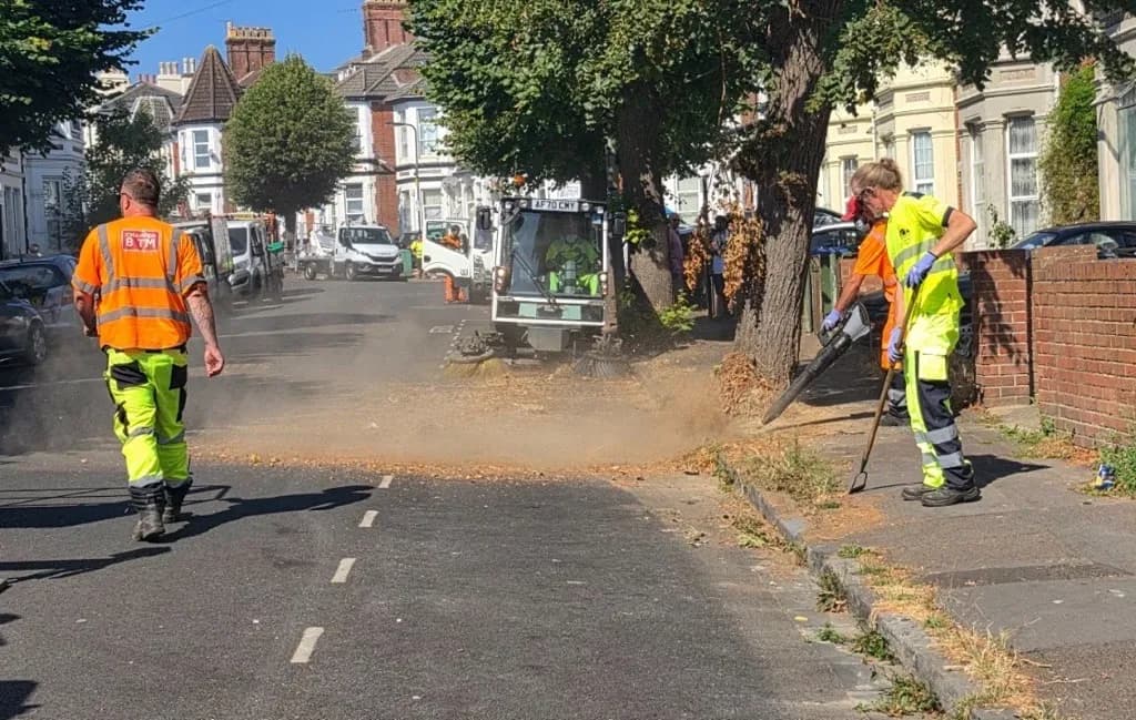 Street sweepers in high-visibility clothing are clearing debris from a residential street with a leaf blower and a street sweeping vehicle.