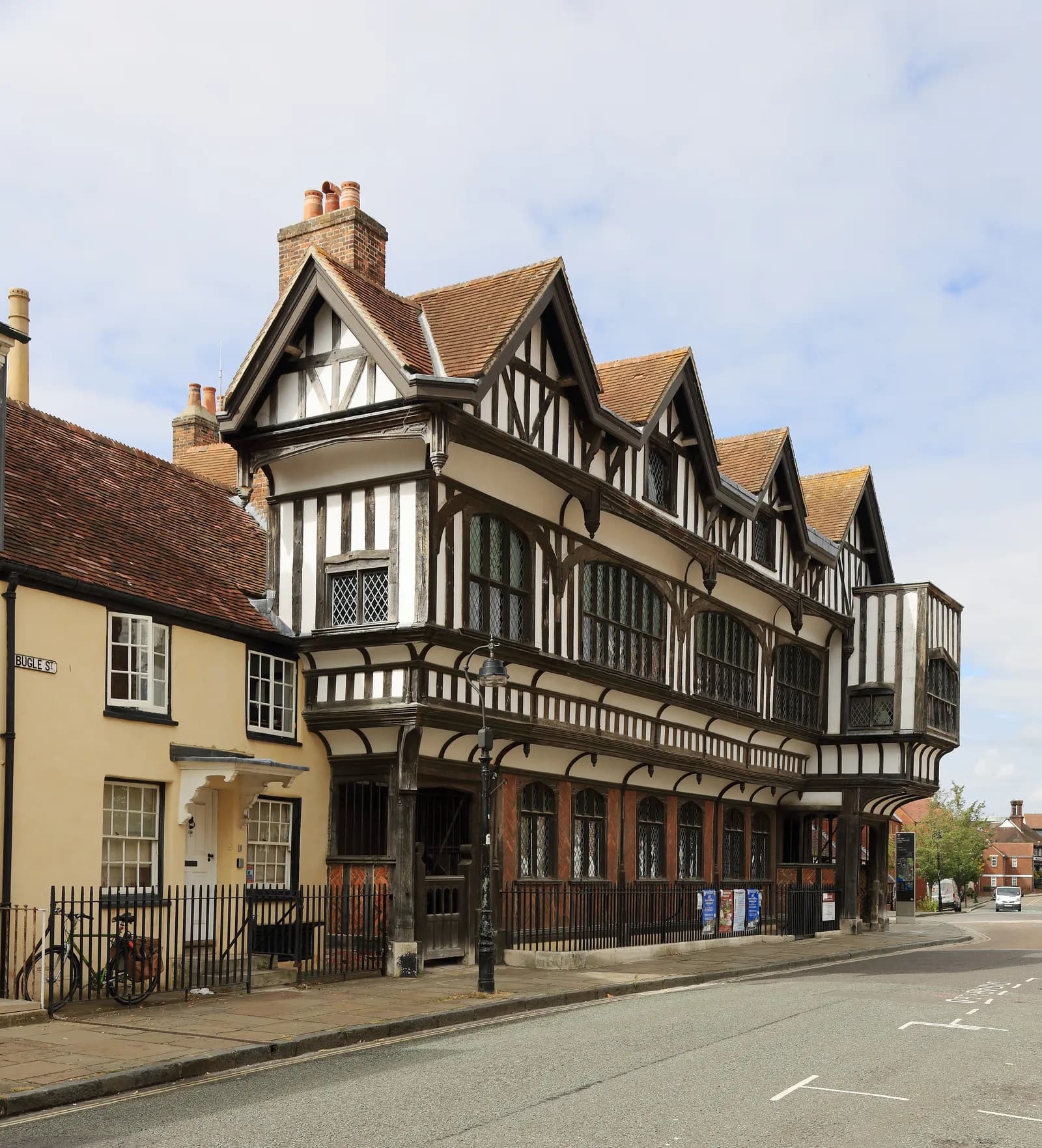 A historic timber-framed building with a white facade and dark brown beams stands prominently next to a lighter yellow building along a paved street.